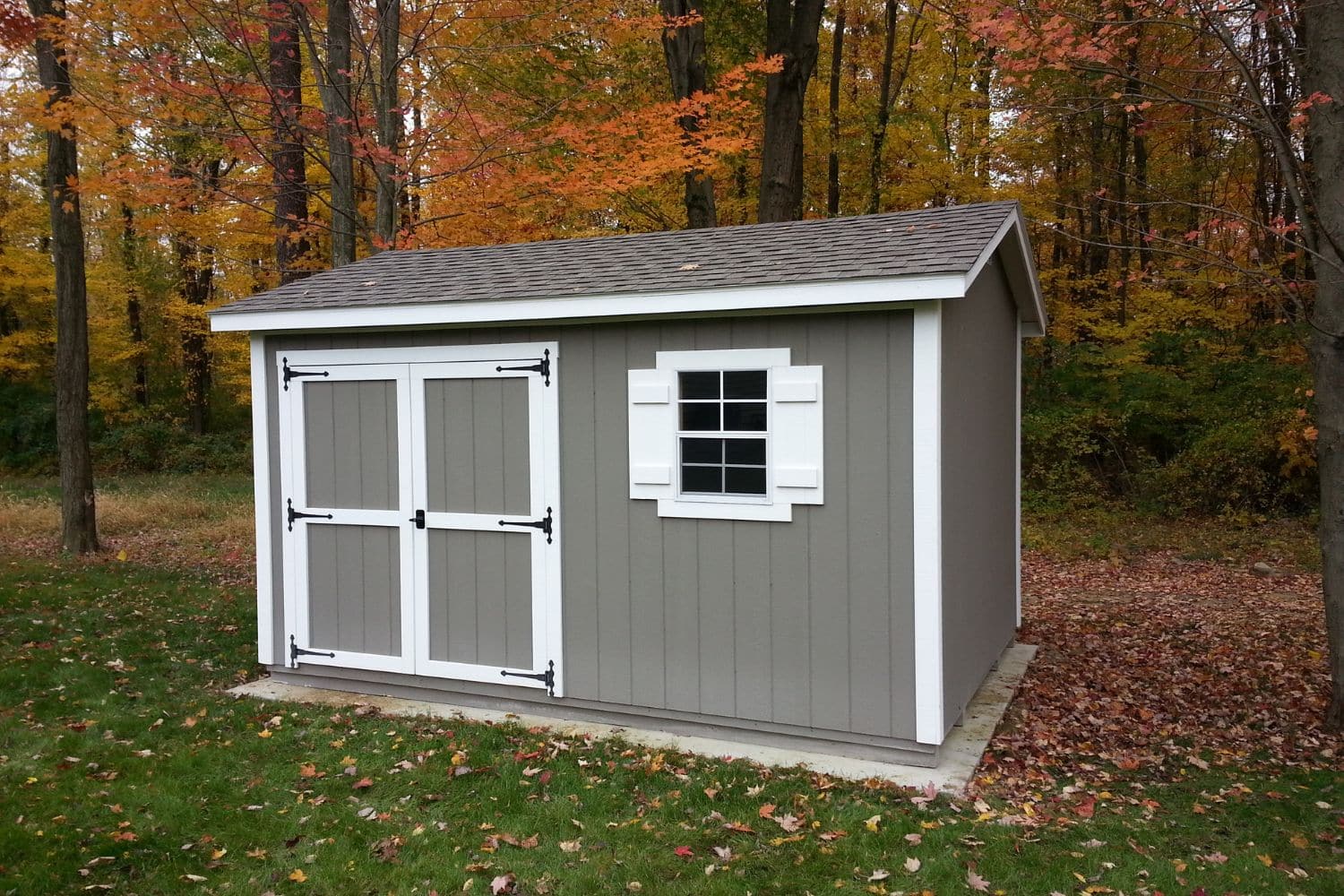 One of our gray and white Gable sheds for sale in Worthington OH - Gray siding with white-trimmed double doors and window beneath gray roof - set near green lawn in front of autumn woods