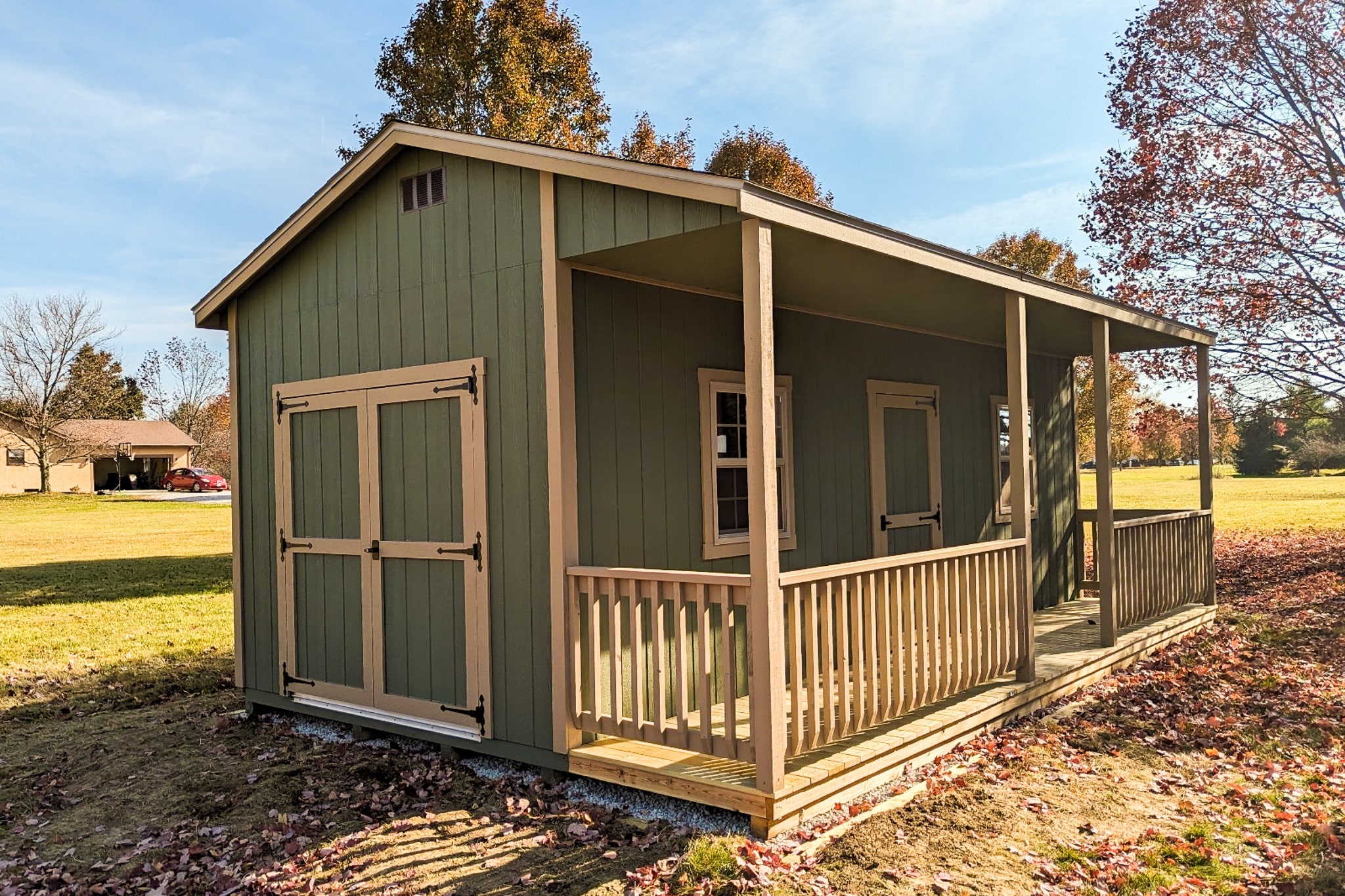 One of our green and beige cabin sheds in Gahanna OH - green wood siding with beige-trimmed double side doors and single front door between two windows overlooking fenced wooden porch with beams supporting overhanging roof - Shed is set on green grass in bed of bronze fallen leaves
