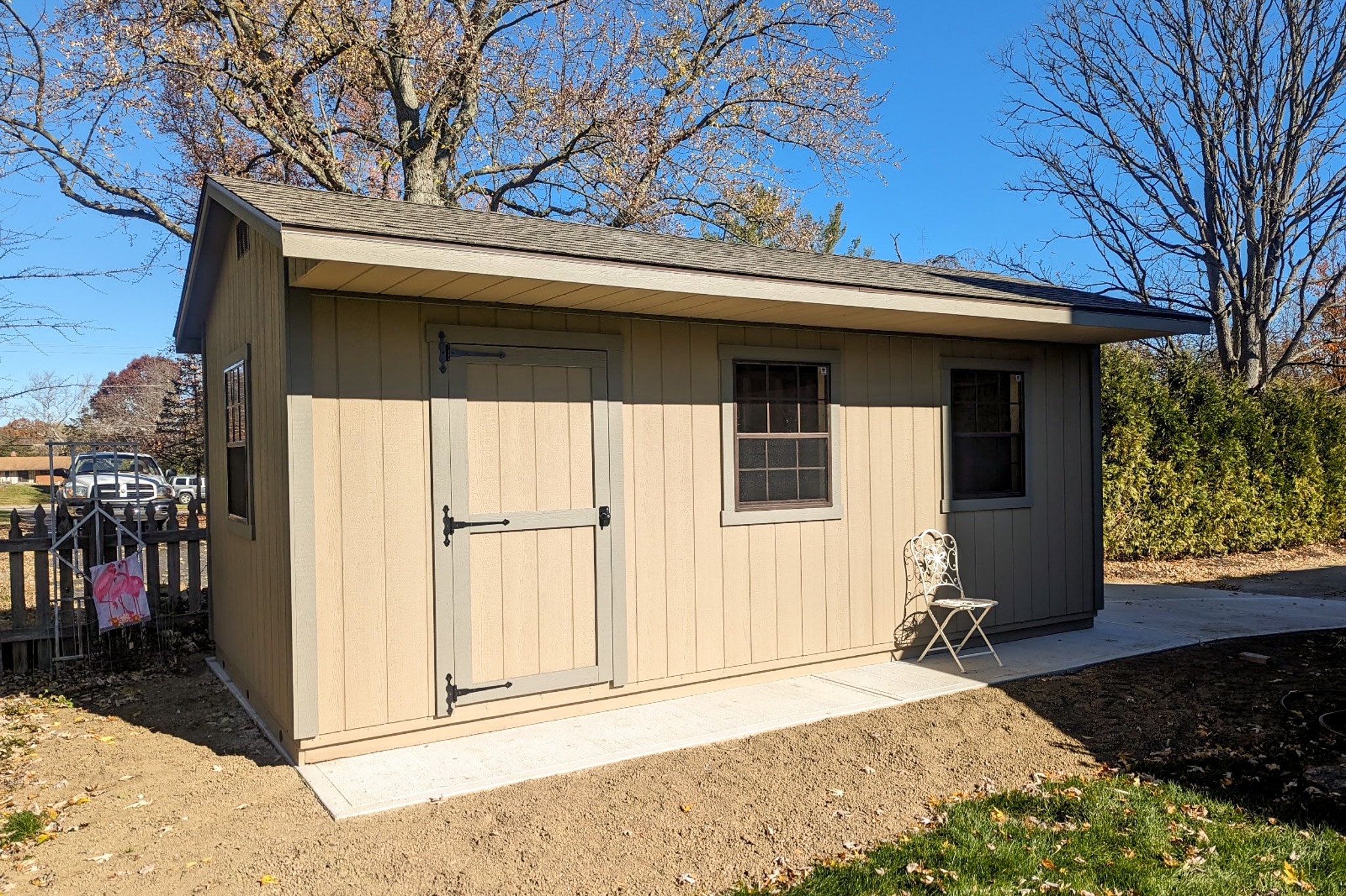 One of our Tan and brown Quaker sheds in Gahanna OH - Tan colored wood siding with khaki colored trimmed door and two windows beside door and one on left side wall - brown shingled roof - shed on property with partially visible adjacent parking lot with light-colored cars - foreground includes grass and dirt ground