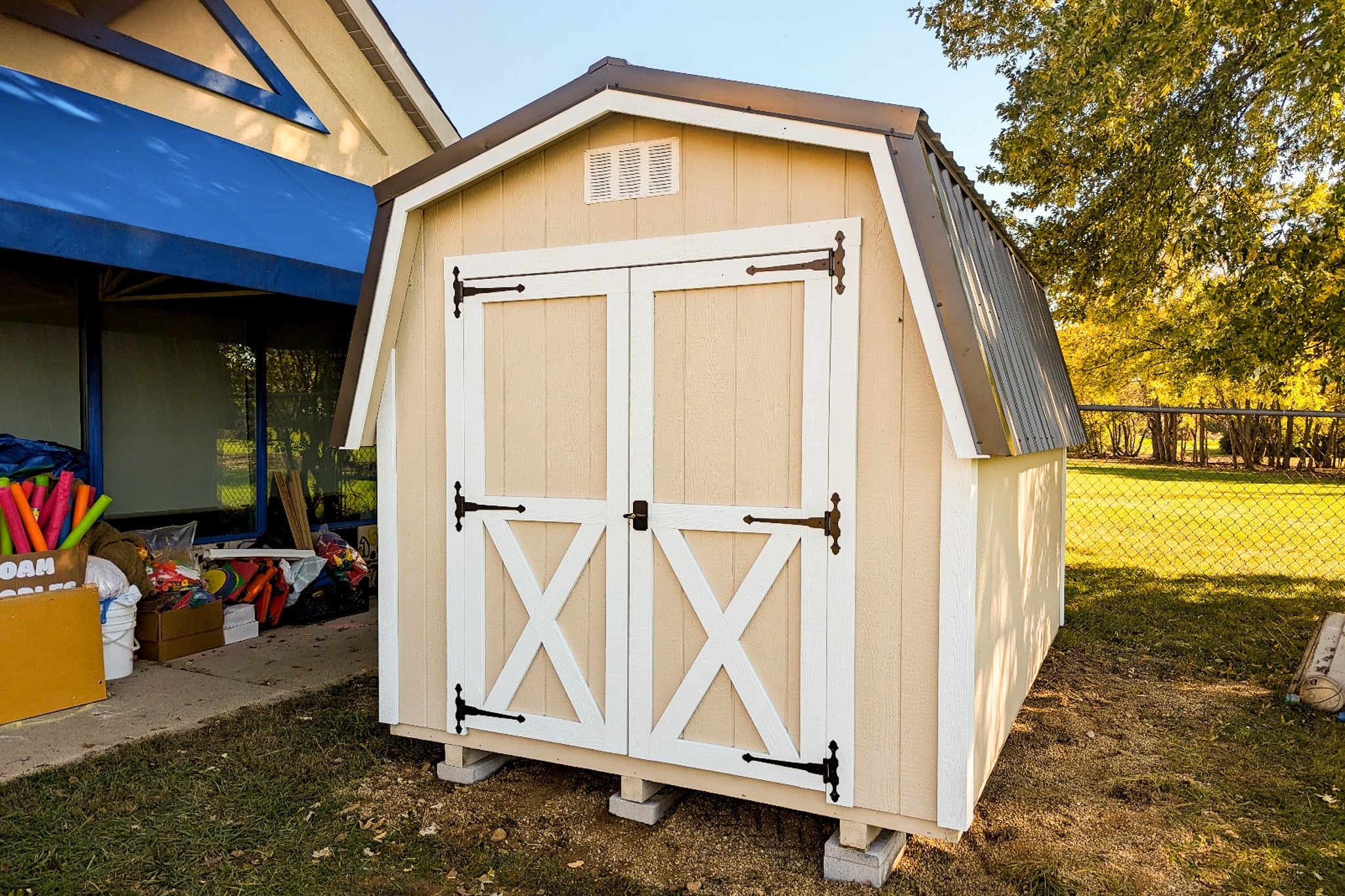 One of our peach and white classic barn-style sheds for sale in Gahanna OH - Peach wooden siding with white trimmed double doors beneath white vent and brown metal roof - Assorted colorful items on the ground to the far left - fenced off grassy tree-lined area in the rear
