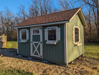 One of our green and beige storage sheds for sale in Lincoln Village OH - Green siding with beige-trimmed windows and door - brown shingled roof - grassy and mulch-covered ground near barren trees