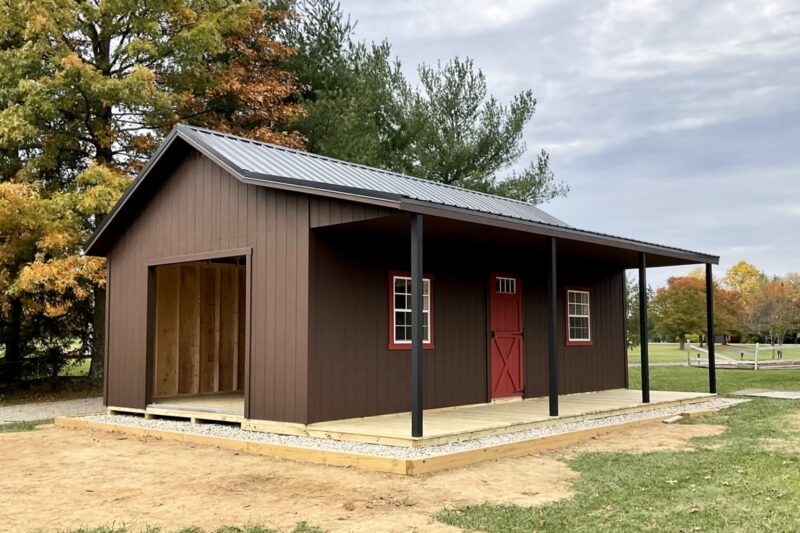 One of our brown retreat sheds for sale in Lincoln Village OH - Brown siding with red and white door and windows - four black beams support black overhanging roof above natural wood porch - set on partially grassy area among trees on cloudy day
