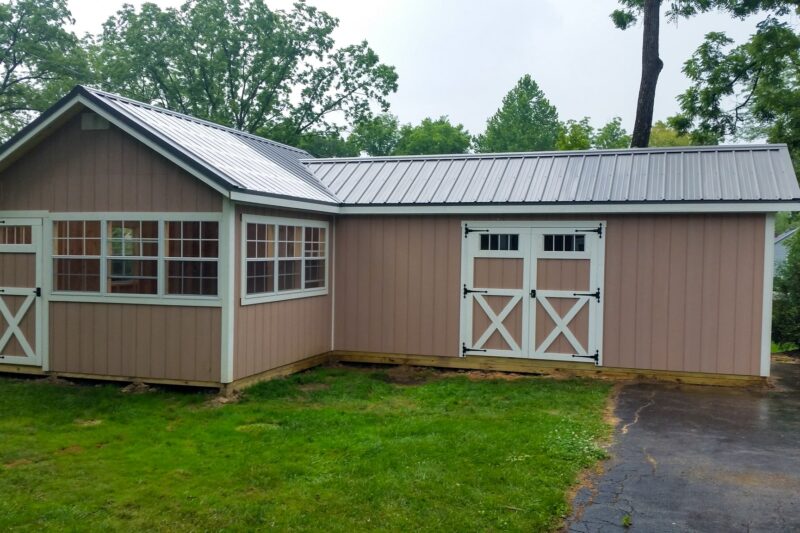 One of our peach and white custom sheds for sale in Lincoln Village OH - Peach L-shaped shed with white-framed double doors on longer side single white-framed windows and door on shorter side under dark gray roof - set on green grass and dark pavement amid trees on cloudy day