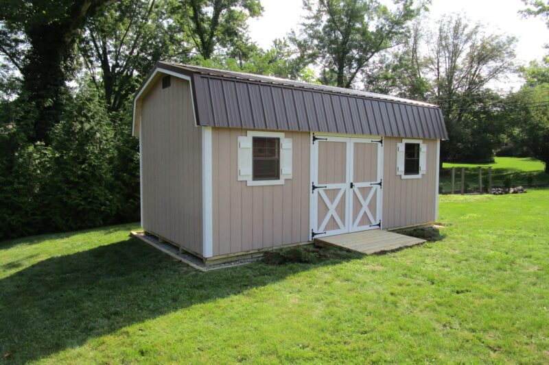 One of our beige Highwall Sheds For Sale Lincoln Village OH - Beige siding with white trimmed double doors and windows with white shutters under brown roof - set on green lawn framed by trees