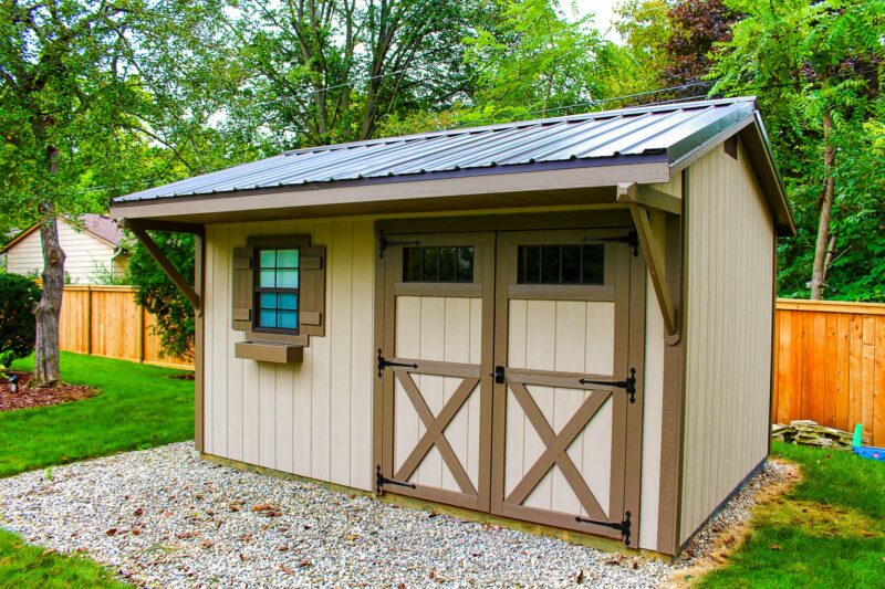 One of our Quaker sheds in Upper Arlington OH - Beige wood siding with brown trimmed double doors and shutters on single window to the left under dark metal roof - shed sits on gravel foundation near green grass and trees in background