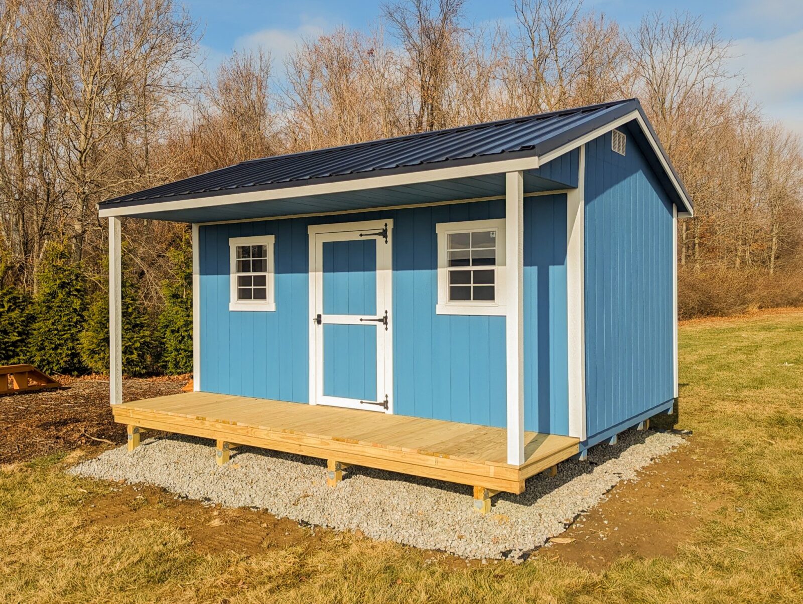 One of our blue and white cabin sheds in Central Ohio - Blue painted wood siding with white trimmed center door flanked by two matching trimmed windows under dark metal roof - White support beams sandwiched between roof and natural wood porch