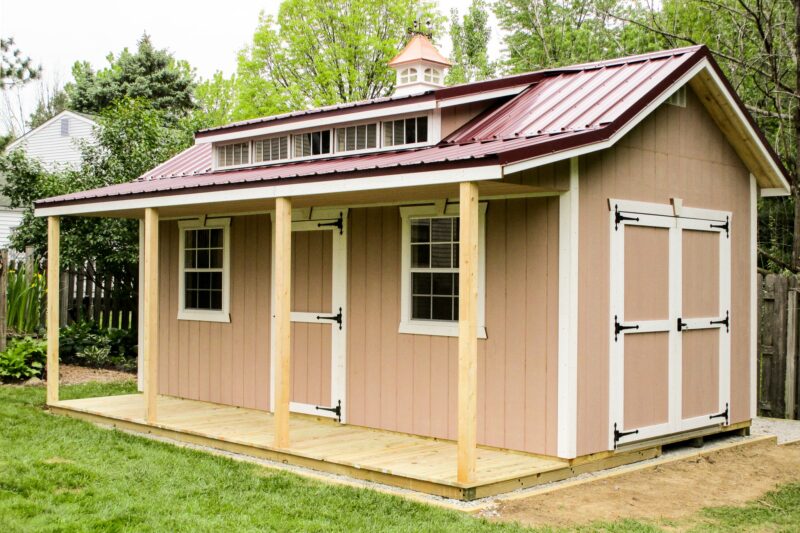 One of our custom sheds in Gahanna OH - Peach siding with white- trimmed double doors on the side - white-trimmed single door between two white-framed windows beneath deep red cupola-topped overhanging roof with loft window - Light wood porch with four matching beams connected to roof - patch of green lawn in foreground and green trees in background