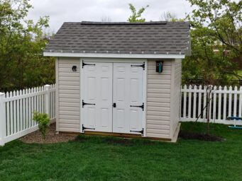 One of our gable sheds in Urbana OH -Beige metal siding with white double doors beneath dark gray shingled roof - shed sits in corner of green grass yard with white picket fence framed by trees