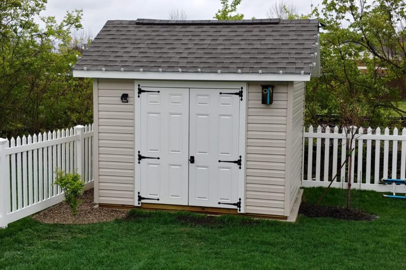 One of our gable sheds in Urbana OH -Beige metal siding with white double doors beneath dark gray shingled roof - shed sits in corner of green grass yard with white picket fence framed by trees