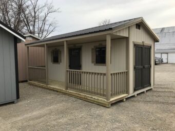 One of our beige cabin sheds in Urbana OH - light beige wood siding with beige trim and matching fenced porch - black double side doors and front black single doors between two windows under black metal roof - shed sits on gravel and dirt lot near other partially visible sheds on cloudy day