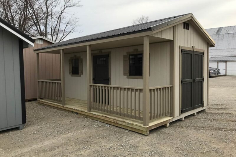 One of our beige cabin sheds in Urbana OH - light beige wood siding with beige trim and matching fenced porch - black double side doors and front black single doors between two windows under black metal roof - shed sits on gravel and dirt lot near other partially visible sheds on cloudy day