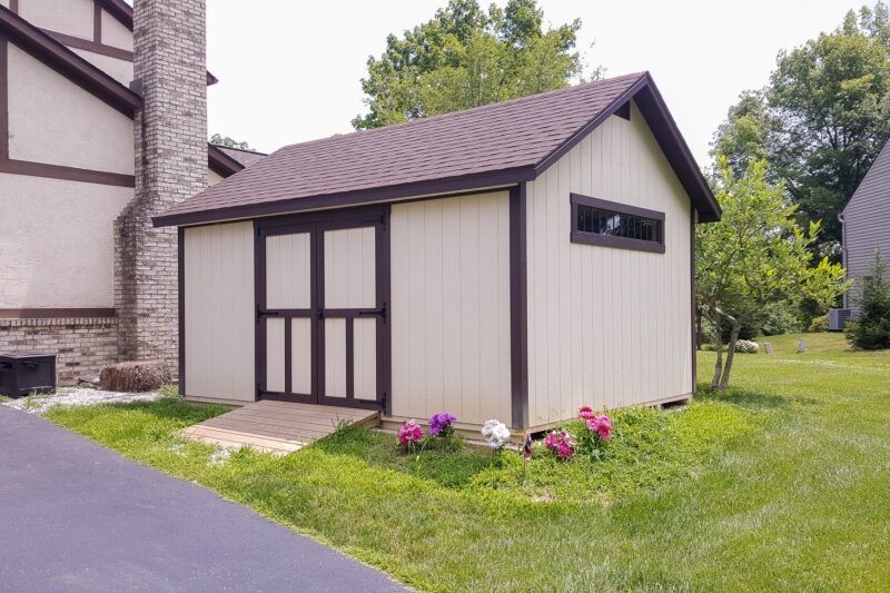 One of our beige and brown Cape Cod sheds in Urbana OH - Beige siding with brown-trimmed double doors and corners - horizontal window on side - brown shingled roof - shed sits on green lawn next to dark paved driveway near beige and brown stucco home with brick chimney