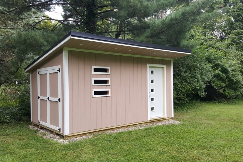 One of our peach and white Custom Sheds in Urbana OH - Peach wood siding with white-trimmed double doors and corners - Three narrow horizontal windows in front with white door inset with 5 small square windows - sloped overhang roof trimmed in black and white - shed sits on grassy yard bordered with green trees