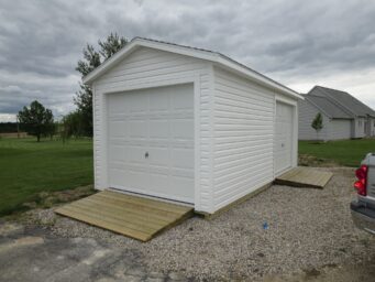 One of our highwall garage sheds in Urbana OH - white siding with two overhead doors in front of light colored wood ramps - sheds sits on gravel in yard near green grass and a few trees - gray house in near distance