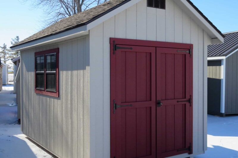 One of our Cape Cod sheds in Urbana OH - Light beige wood siding with red-trimmed red double doors and red-trimmed side windows under dark brown shingled roof