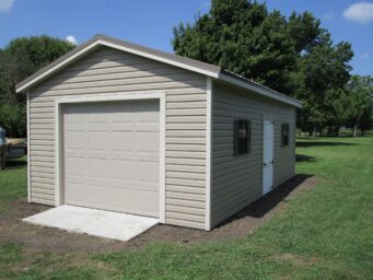 One of our beige garage sheds in Urbana OH - Beige metal siding with beige overhead door with white side door between two windows with brown shutters beneath beige metal roof