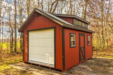 One of our red cottage sheds in Whitehall OH - Red wood siding with black-trimmed corners and single side door between two matching windows - black-framed white overhead door beneath black metal roof - row of window panes adorn loft area - shed sits beside trees with sparse autumn leaves